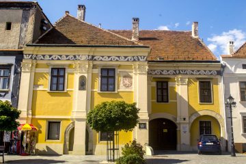 Yellow building with brown roof, arched doorway, and blue car parked in front on a sunny day.
