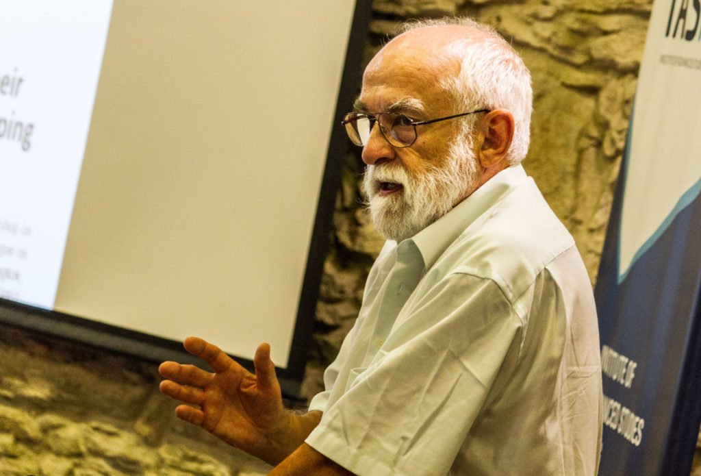 Idős férfi szemüvegben, fehér szakállal, előadást tart az iASK nyári egyetemén.