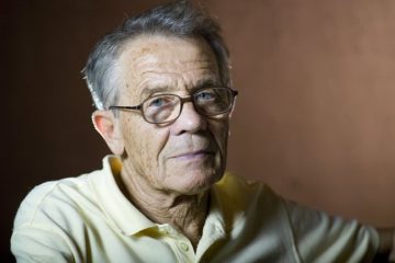 Idős férfi szemüvegben, sárga pólóban néz a kamerába.