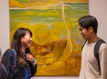 Két ember mosolyog Mihály Magdolna kiállításán, színes festmény a háttérben.