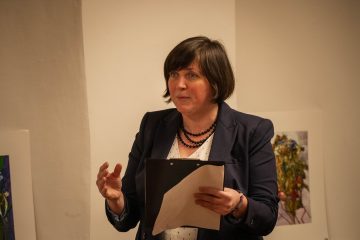 Mihály Magdolna kiállításán egy nő előadást tart, kezében papírokkal.