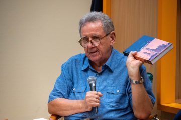 Idősebb szemüveges férfi mikrofonba beszél, kezében Csillogás és katasztrófa könyv