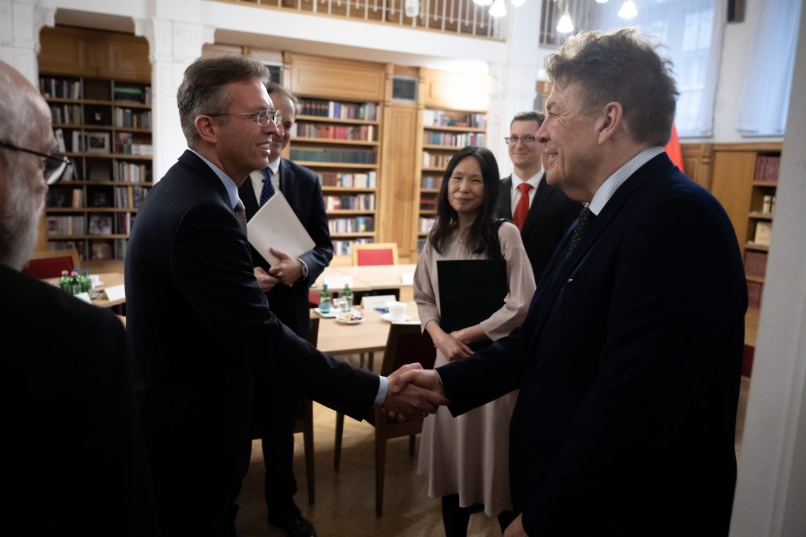 Két férfi kezet fog Kőszegen, a háttérben könyvespolcok és más emberek láthatók.