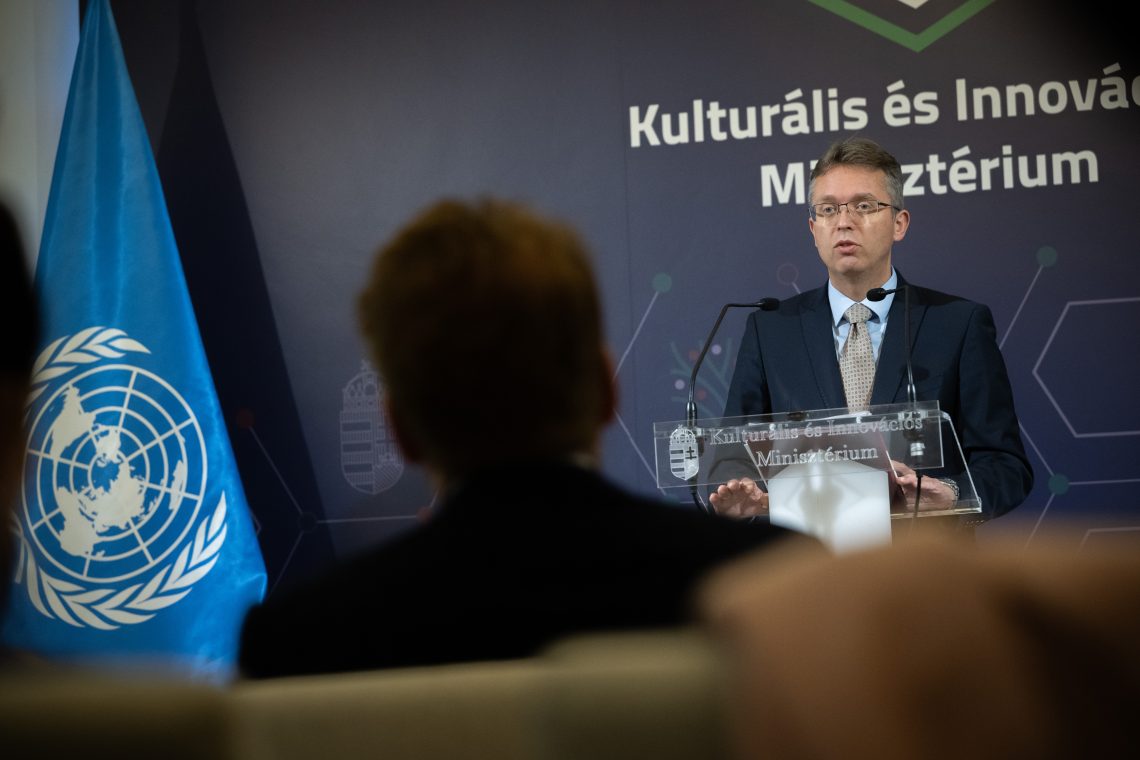 Férfi beszél a pódiumon, ENSZ zászló a háttérben. Kulturális és Innovációs Minisztérium.