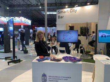 Nő a "Smart City Bar"-nál a barcelonai iASK Smart City Expo Világkongresszuson. Magyar Kereskedelmi és Iparkamara logó.