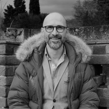 Mosolygó, szemüveges férfi télikabátban egy téglafal előtt.